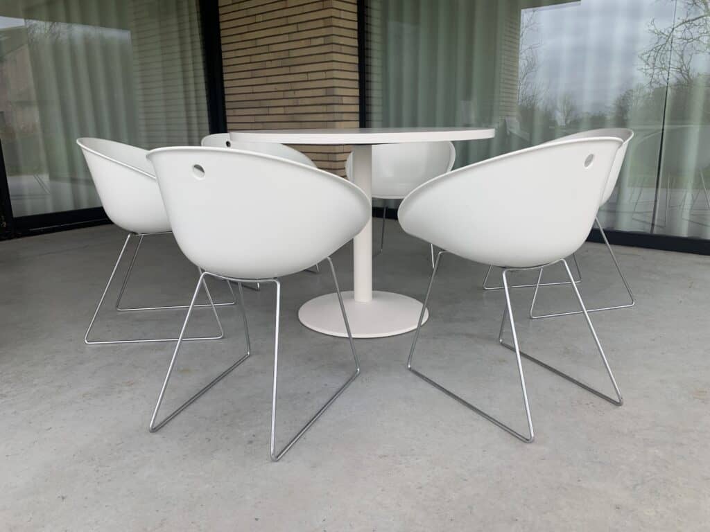 Vijf moderne witte stoelen met metalen poten staan rond een ronde witte tafel op een grijze betonnen vloer, voor grote glazen ramen met gesloten witte gordijnen.