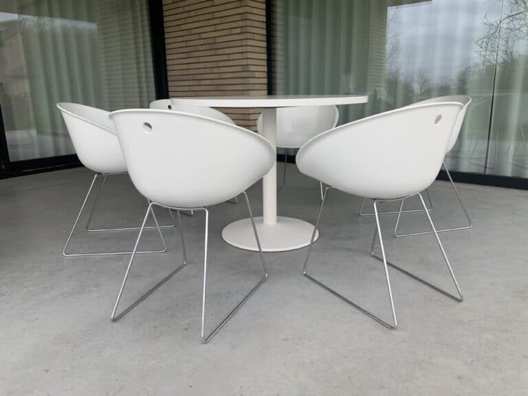 Vijf moderne witte stoelen met metalen poten staan rond een ronde witte tafel op een grijze betonnen vloer, voor grote glazen ramen met gesloten witte gordijnen.