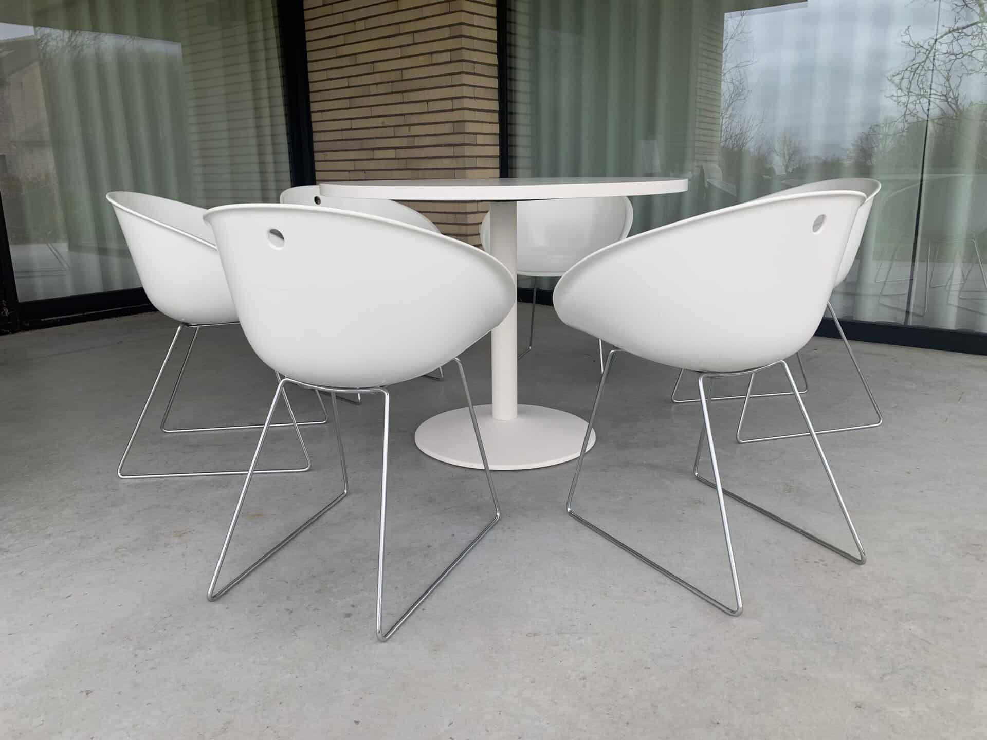Vijf moderne witte stoelen met metalen poten staan rond een ronde witte tafel op een grijze betonnen vloer, voor grote glazen ramen met gesloten witte gordijnen.