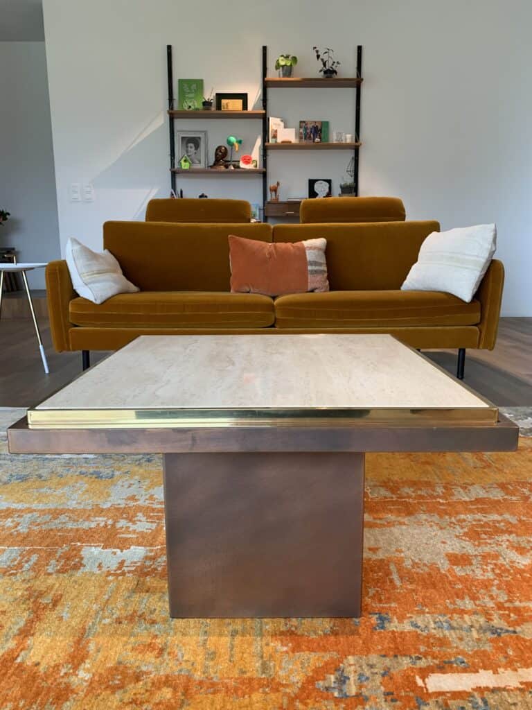 Een moderne woonkamer met een mosterdgele bank, witte en oranje kussens, een salontafel met marmeren blad en bronzen onderstel en een kleurrijk oranje vloerkleed. Aan de muur achter de bank hangen planken met boeken en decoratie.