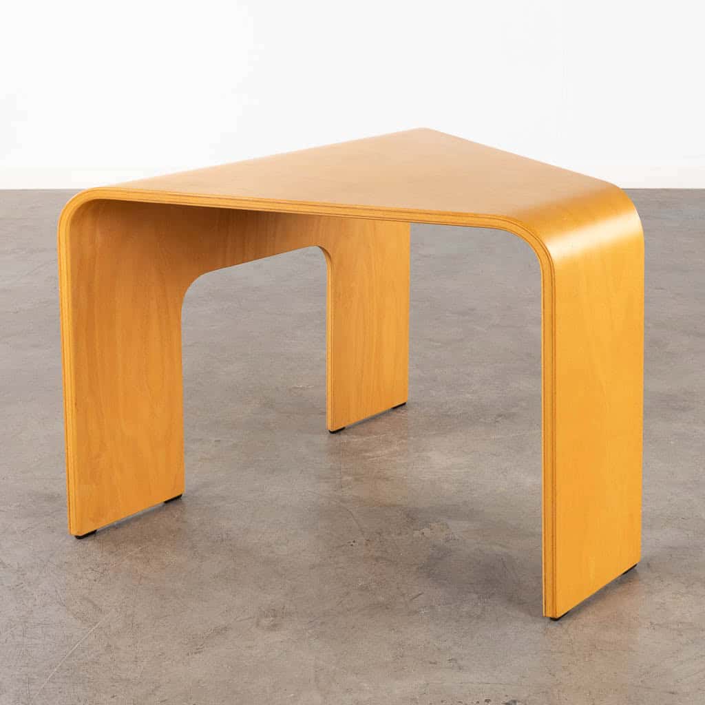 Een minimalistische, driehoekige houten tafel met afgeronde hoeken en gladde oppervlakken, geplaatst op een grijze betonnen vloer tegen een eenvoudige witte muur.