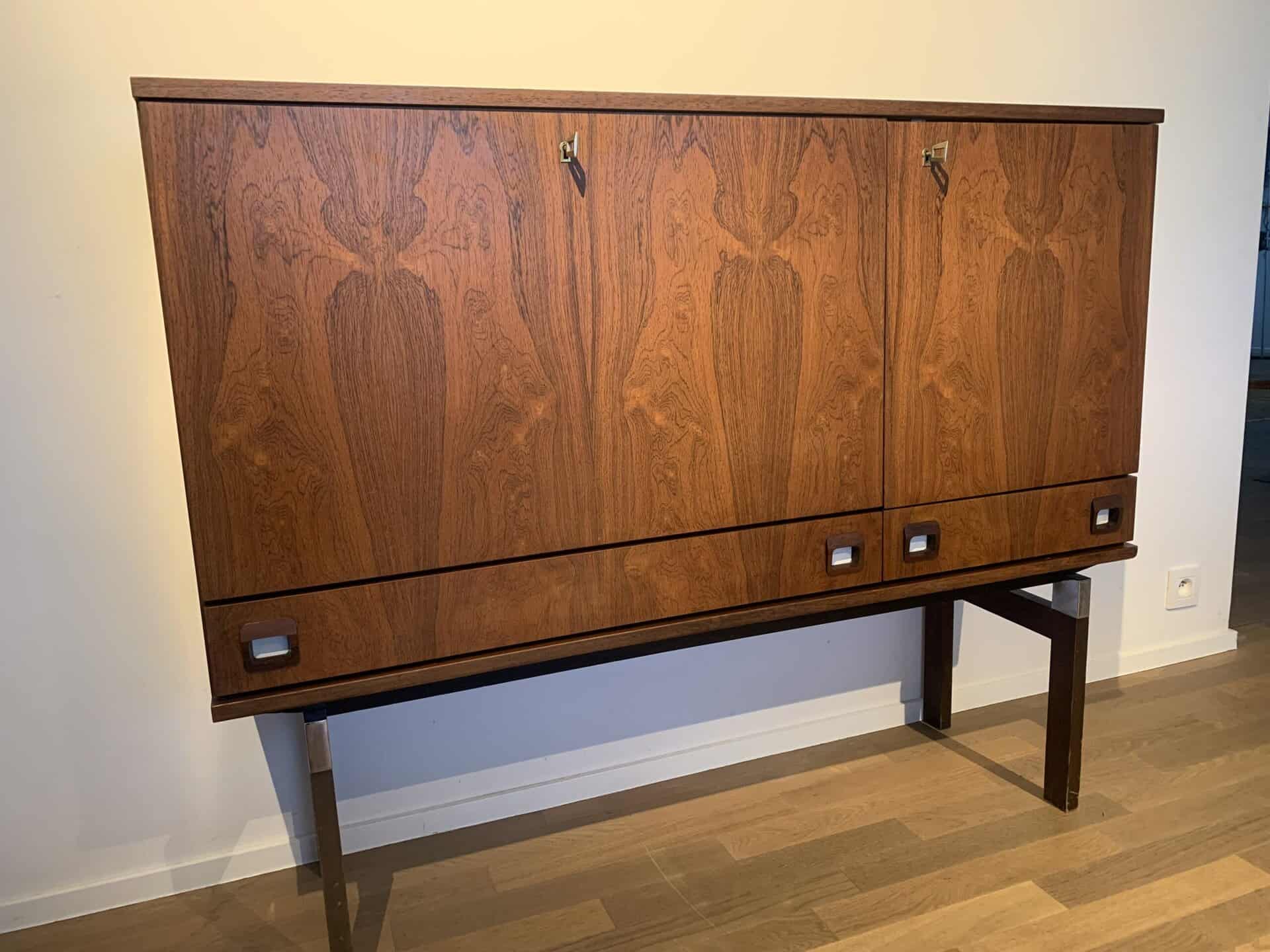 Een vintage houten kast met drie deuren met een symmetrisch houtnerfpatroon, metalen handgrepen en slanke poten, staat tegen een lichtgekleurde muur op een houten vloer.