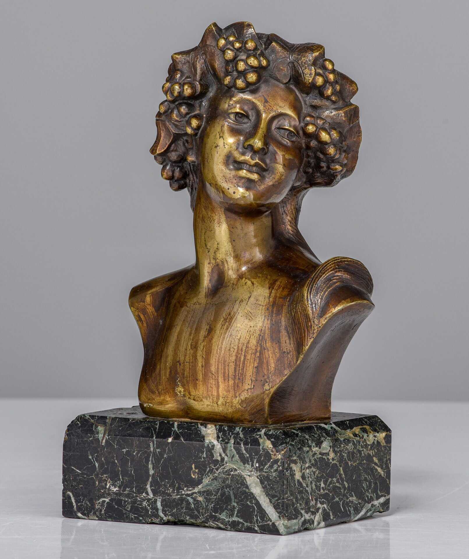 Een bronzen buste van een vrouw met druiventrossen en bladeren in haar haar, gemonteerd op een zwarte marmeren sokkel, tegen een effen grijze achtergrond.