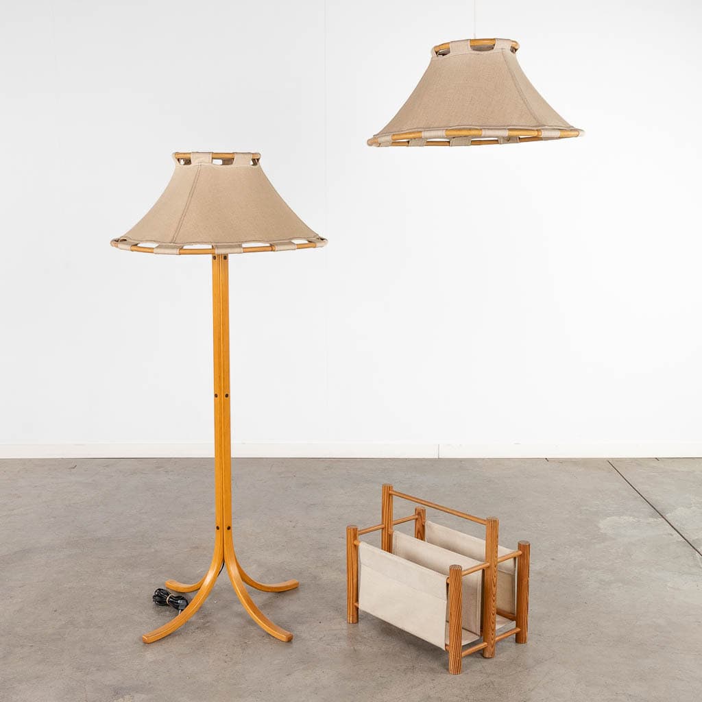Een houten staande lamp met een beige stoffen kap, een bijpassende hanglamp en een klein houten tijdschriftenrek met beige stof, alles tegen een eenvoudige witte muur en betonnen vloer.