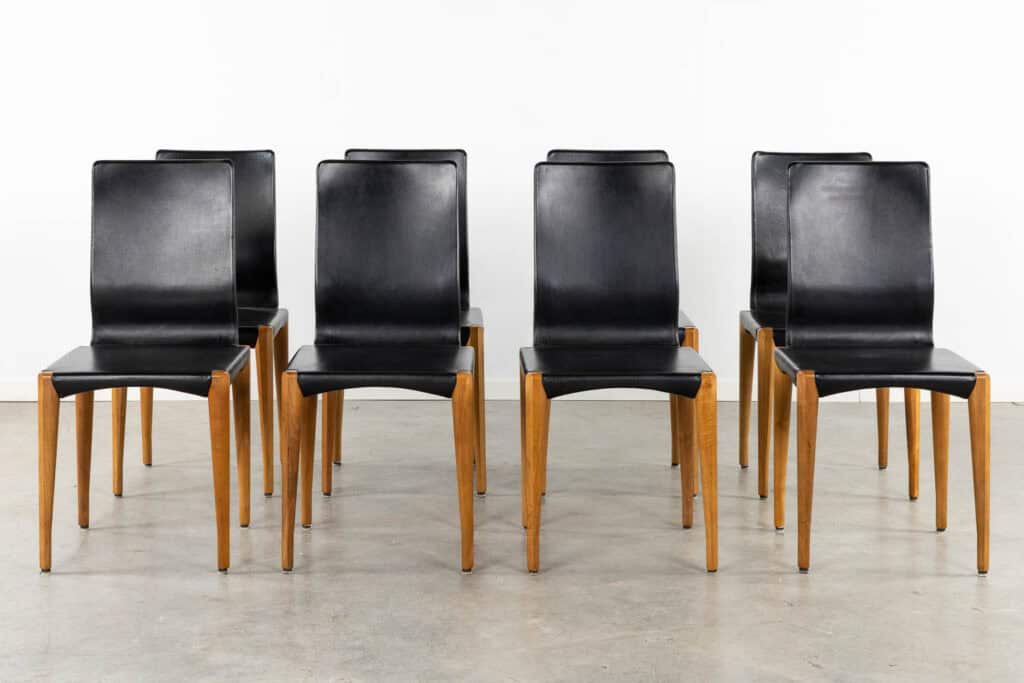Zes moderne eetkamerstoelen met zwarte leren zittingen en rugleuningen en lichte houten poten, gerangschikt in twee rijen op een lichtgrijze betonnen vloer tegen een eenvoudige witte muur.