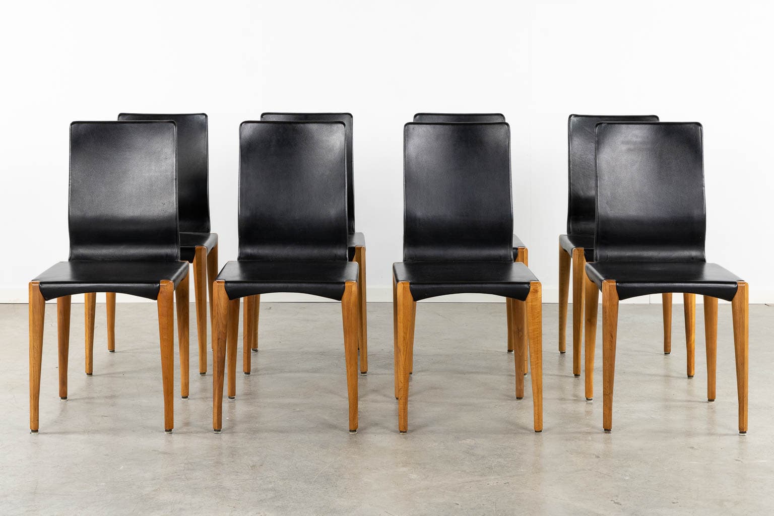Zes moderne eetkamerstoelen met zwarte leren zittingen en rugleuningen en lichte houten poten, gerangschikt in twee rijen op een lichtgrijze betonnen vloer tegen een eenvoudige witte muur.