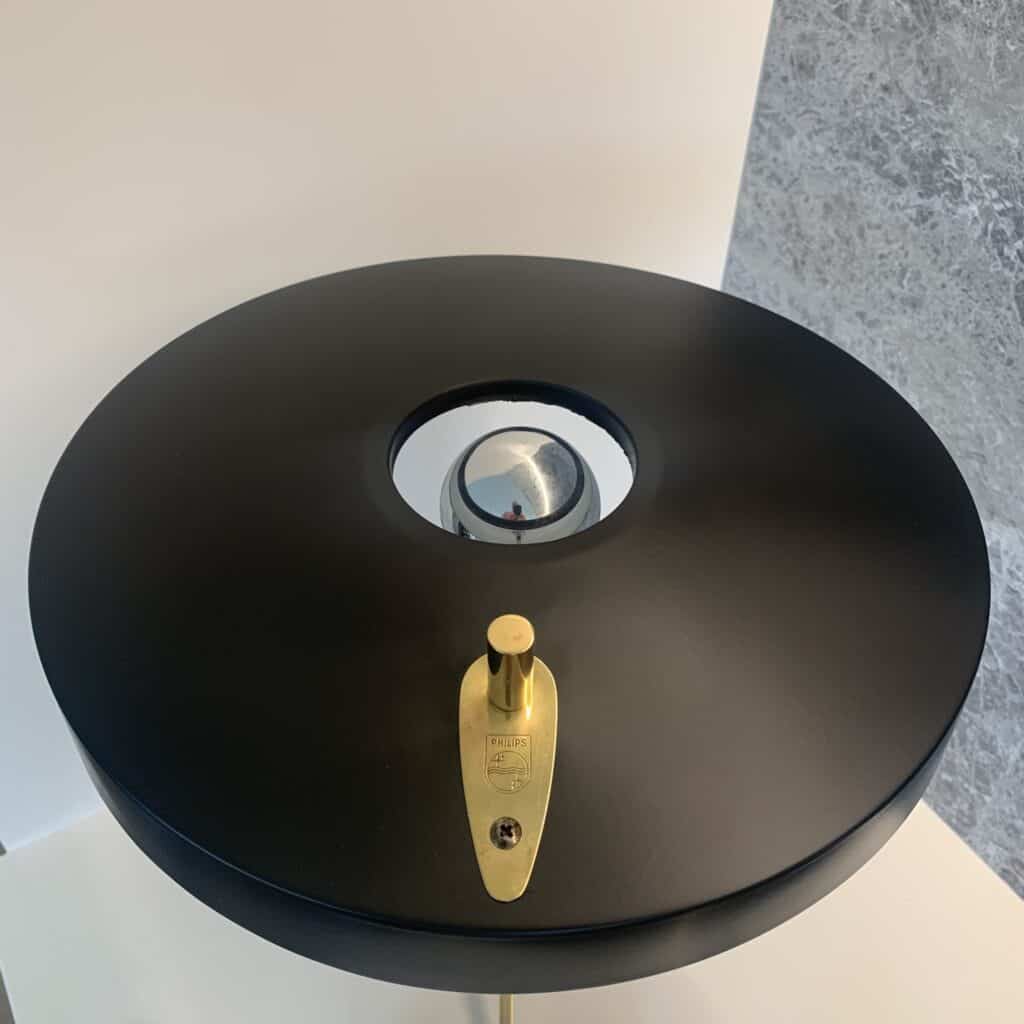 Een close-up van een moderne, ronde, zwarte lamp met een metalen messing schakelplaat en knop en een reflecterende zilveren bol in het midden, tegen een wit-grijze achtergrond.