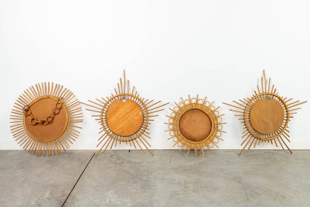 Vier ronde wandspiegels of -schalen in sunburst-stijl met complexe houten lijsten staan opgesteld tegen een witte muur op een betonnen vloer. Elk stuk heeft een uniek radiaal ontwerp met verschillende patronen en vormen.