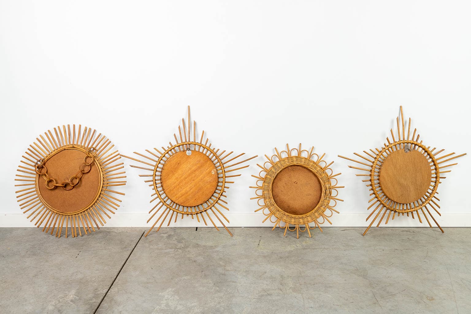 Vier ronde wandspiegels of -schalen in sunburst-stijl met complexe houten lijsten staan opgesteld tegen een witte muur op een betonnen vloer. Elk stuk heeft een uniek radiaal ontwerp met verschillende patronen en vormen.