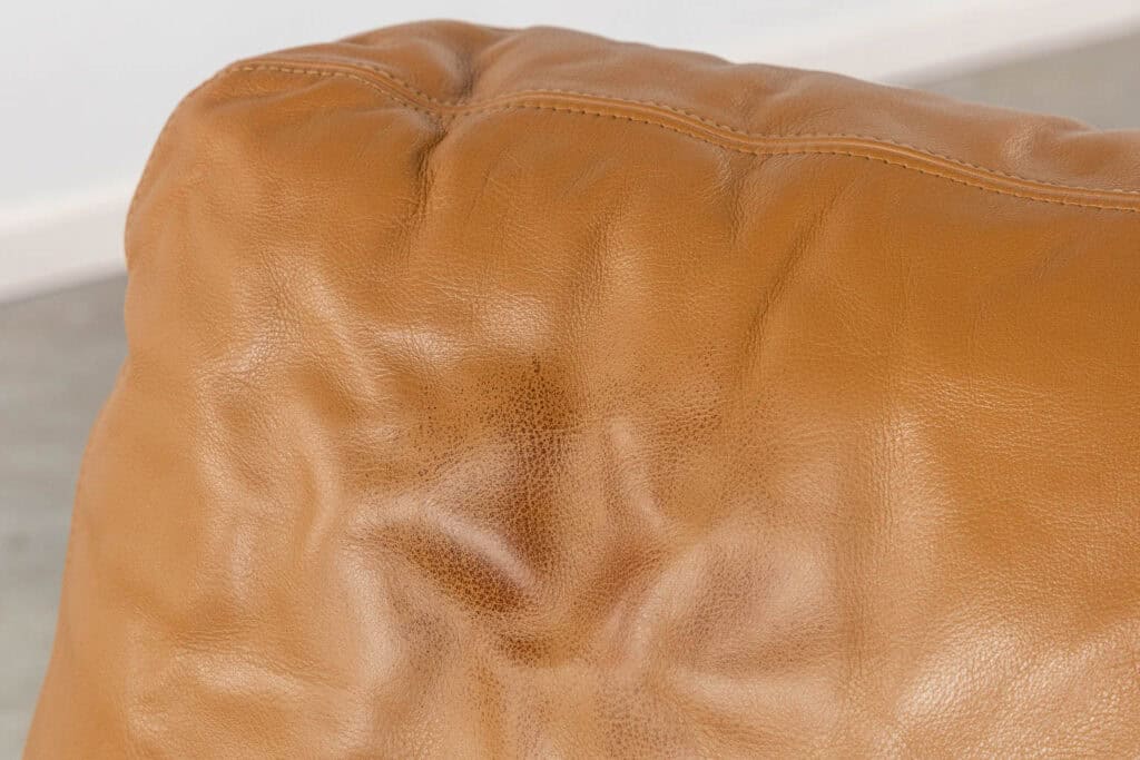 Close-up van een bruin leren kussen met zichtbare stiksels en natuurlijke rimpels, die de textuur en lichte glans van het materiaal laten zien. De achtergrond is wazig.