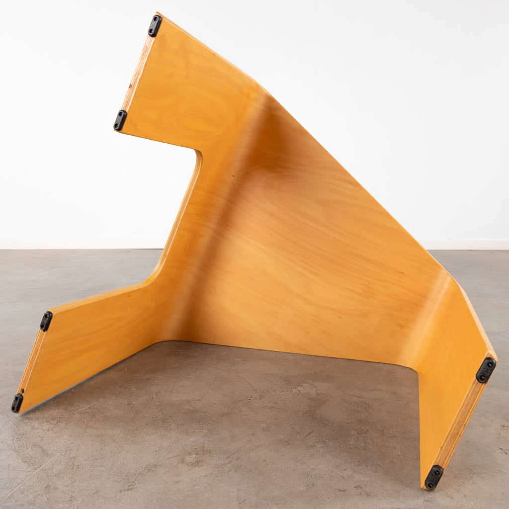 Een moderne, hoekige houten stoel met een sculpturaal ontwerp en rubberen voetjes staat op een gladde betonnen vloer tegen een eenvoudige witte muur.