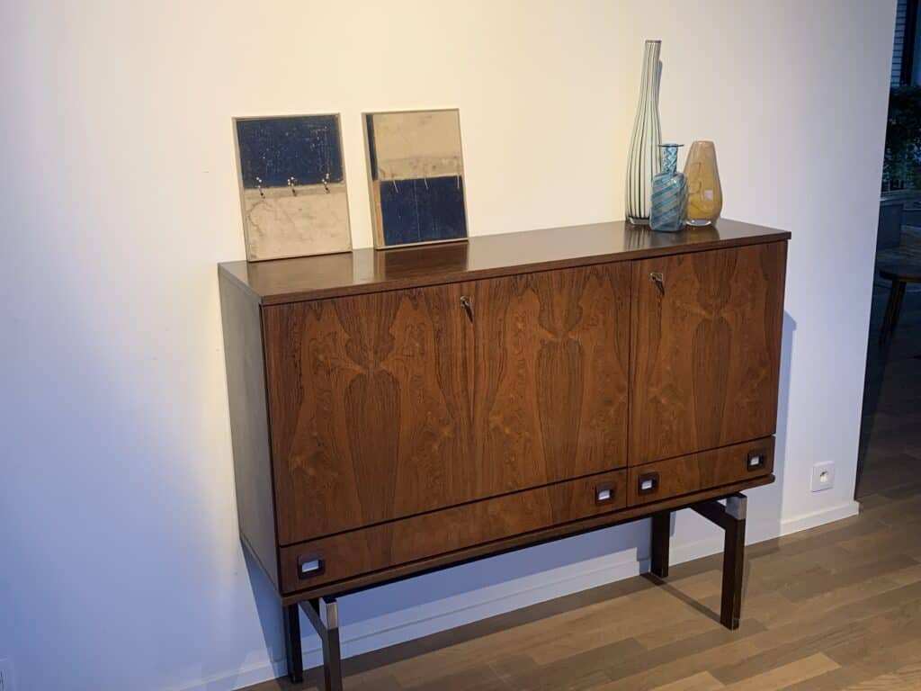 Een houten kast uit het midden van de vorige eeuw met decoratief fineer staat tegen een witte muur. Bovenop staan twee rechthoekige spiegels, een hoge gestreepte vaas, een vaas van helder glas en een vaas van gelig glas. De vloer is van hout.