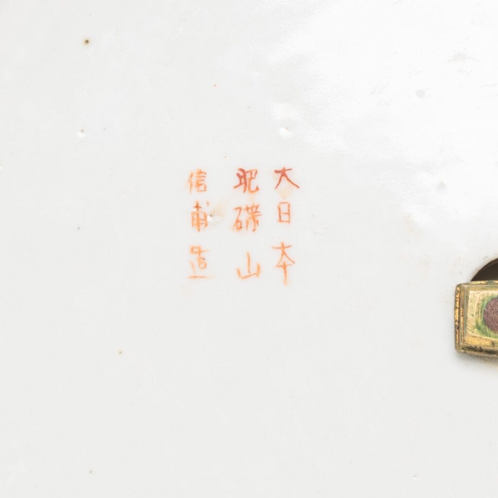 Rode Japanse kanji-tekens zijn geschilderd op een wit keramisch oppervlak, waarschijnlijk een meesterteken. Een klein metalen plaatje is rechtsonder aan de afbeelding bevestigd.
