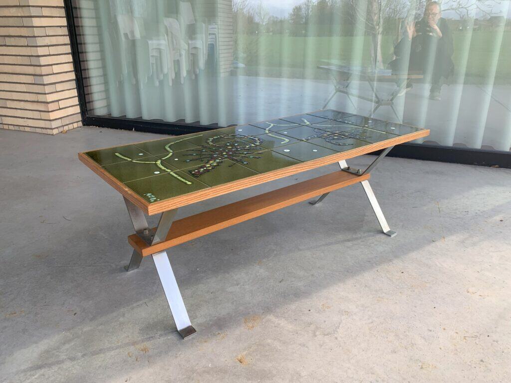 Een houten en metalen bank met een tafelblad met een gedetailleerd kaartontwerp staat op een betonnen vloer buiten een gebouw met glazen deuren. Reflecties in het glas tonen een persoon die de foto neemt en groen gras buiten.