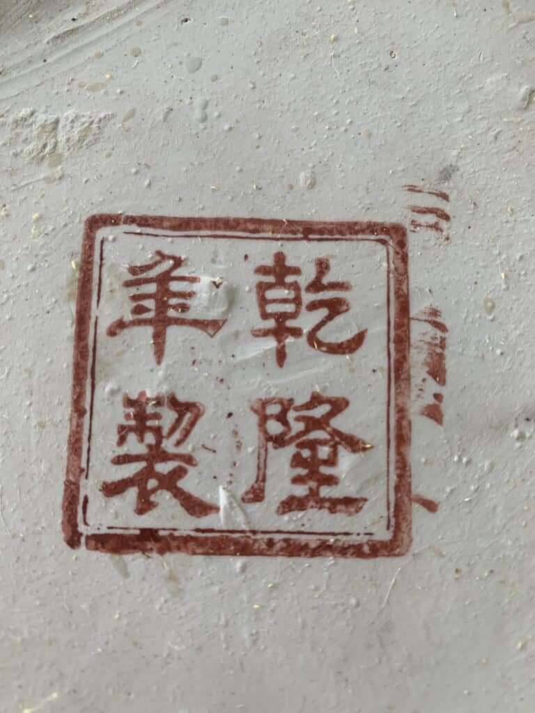 Rode Chinese zegelstempel met vier karakters op een getextureerd wit oppervlak, mogelijk een meesterteken of signatuur op aardewerk of porselein. De karakters zijn in een vierkant geplaatst en omrand met een rode rand.
