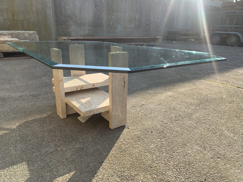 Een moderne salontafel met een dik, rechthoekig glazen blad en een geometrisch stenen onderstel, buiten tentoongesteld op een betonnen ondergrond in het felle zonlicht.