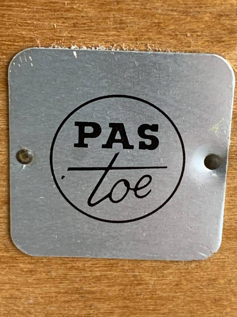 Een metalen plaatje met de tekst “PAS toe” in het zwart, met een ronde rand, met twee schroeven bevestigd aan een houten oppervlak.