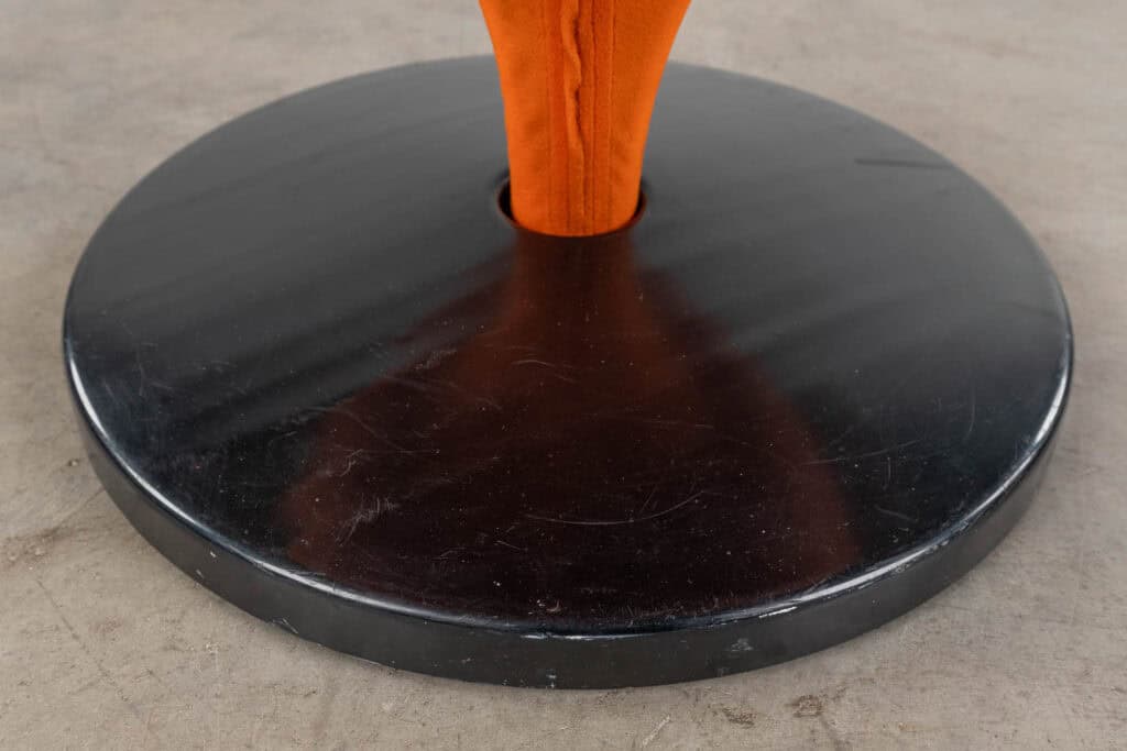 Close-up van een ronde, zwarte basis met in het midden een oranje, met stof beklede paal, geplaatst op een betonnen vloer.