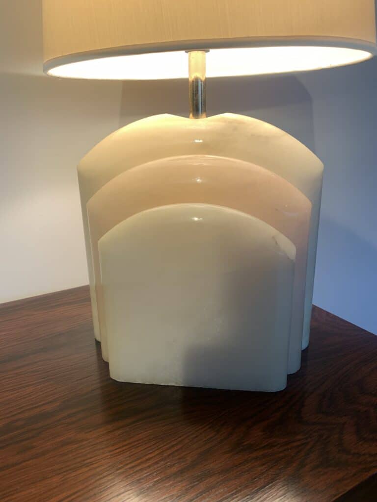Een lamp met een beige kap en een gebeeldhouwde marmeren voet met drie boogvormige lagen, geplaatst op een donkere houten tafel tegen een witte muur.
