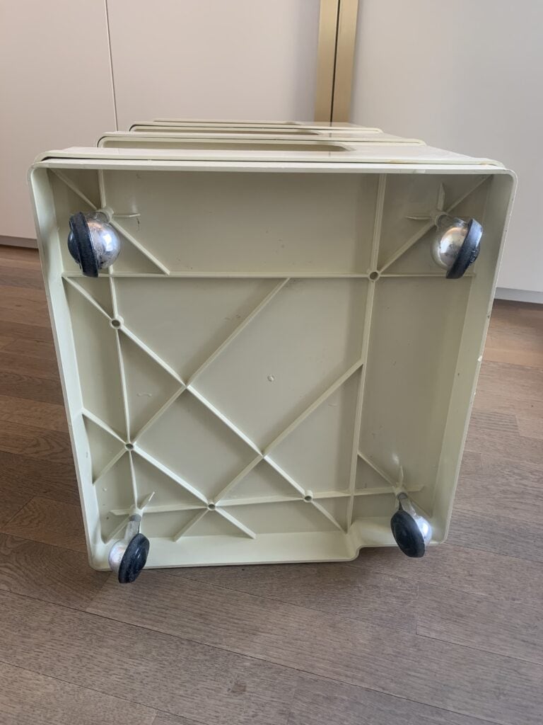 Een blik op de onderkant van een lichtbeige plastic kar met vier zwart-zilveren zwenkwielen, rustend op een houten vloer vlak bij witte kasten.