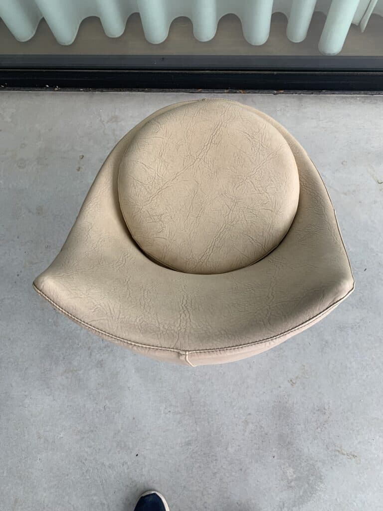 Een beige, ronde, moderne stoel met een gebogen rugleuning staat op een lichtgrijze betonnen vloer. Aan de onderkant van de afbeelding is een deel van iemands donkere schoen zichtbaar. Verticale jaloezieën zijn op de achtergrond te zien.