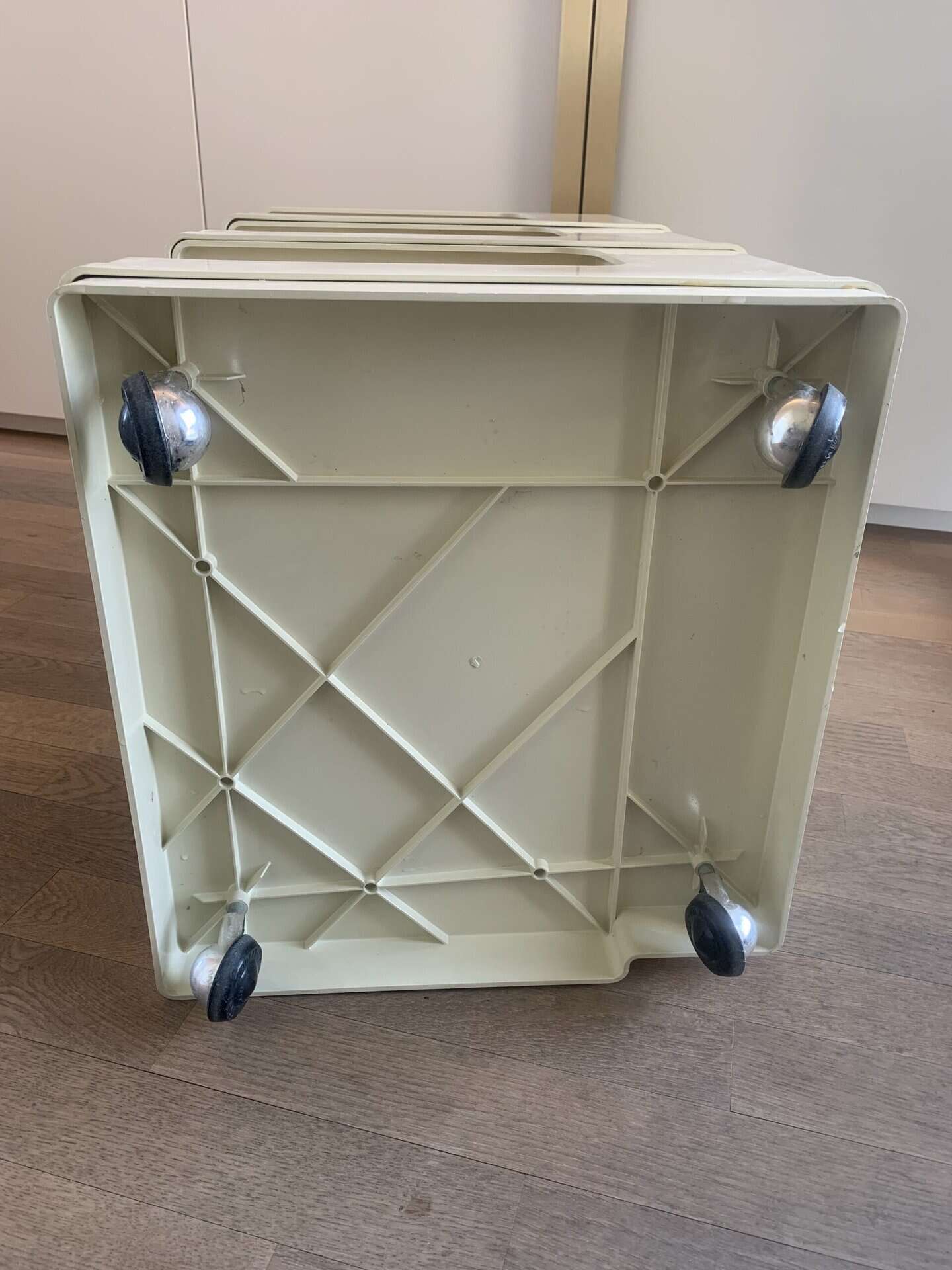 Een blik op de onderkant van een lichtbeige plastic kar met vier zwart-zilveren zwenkwielen, rustend op een houten vloer vlak bij witte kasten.