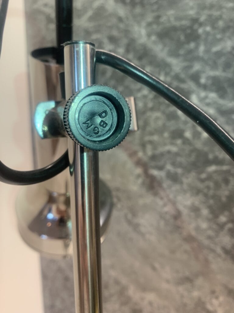 Close-up van een metalen standaard met een zwarte verstelknop met de labels 