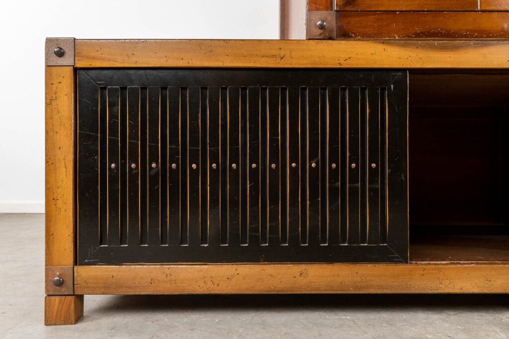 Een close-up van een houten kast met een zwart lamellenpaneel met verticale groeven en ronde metalen accenten, omlijst door verweerd lichtbruin hout met zichtbare verbindingen en metalen hoekdetails.