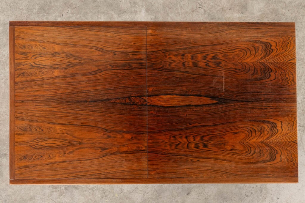 Een rechthoekig houten tafelblad met rijke, donkerbruine houtnerfpatronen en een zichtbare naad in het midden, tegen een grijze betonnen achtergrond.