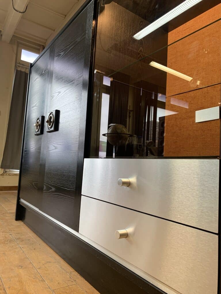 Een moderne kast met zwarte houten deuren, metalen handgrepen, twee witte lades met zilveren knoppen en een glazen displaygedeelte staat op een houten vloer in een goed verlichte kamer.