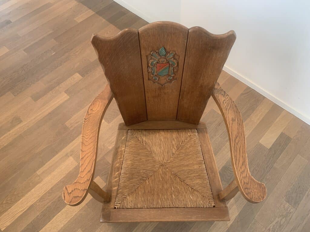 Een houten stoel met een gevlochten zitting en gebogen armleuningen, met een decoratief wapen met een schild en op de rugleuning gesneden en geschilderde bladeren, geplaatst op een lichte houten vloer bij een witte muur.