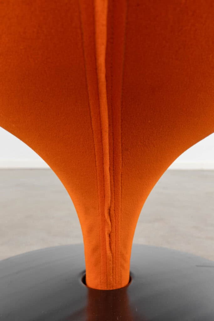 Close-up van een oranje, gebogen gestoffeerde stoelleuning met een naad in het midden, die taps toeloopt in een ronde, zwarte basis op een lichtgrijze vloer.