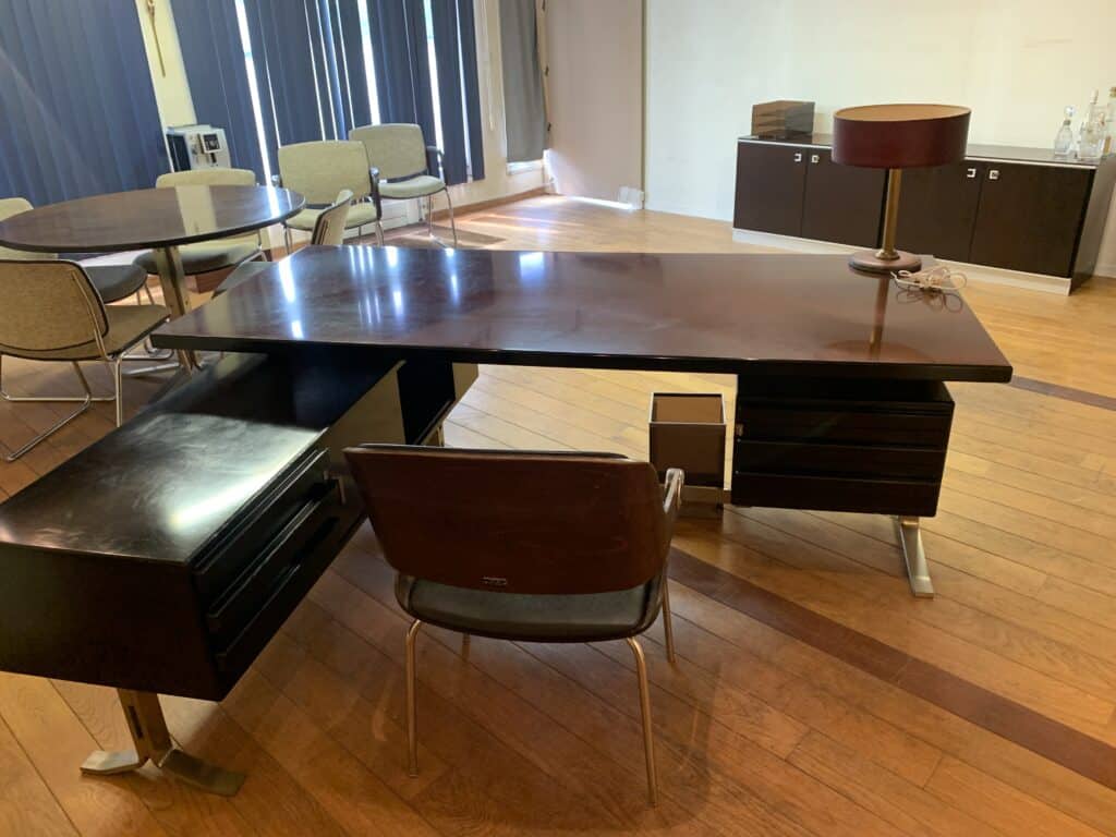Een modern kantoor met een donkerhouten bureau, stoel, lamp en een bril. Er staat ook een ronde tafel met stoelen, een kast en een houten vloer. Natuurlijk licht komt binnen via verticale jaloezieën.