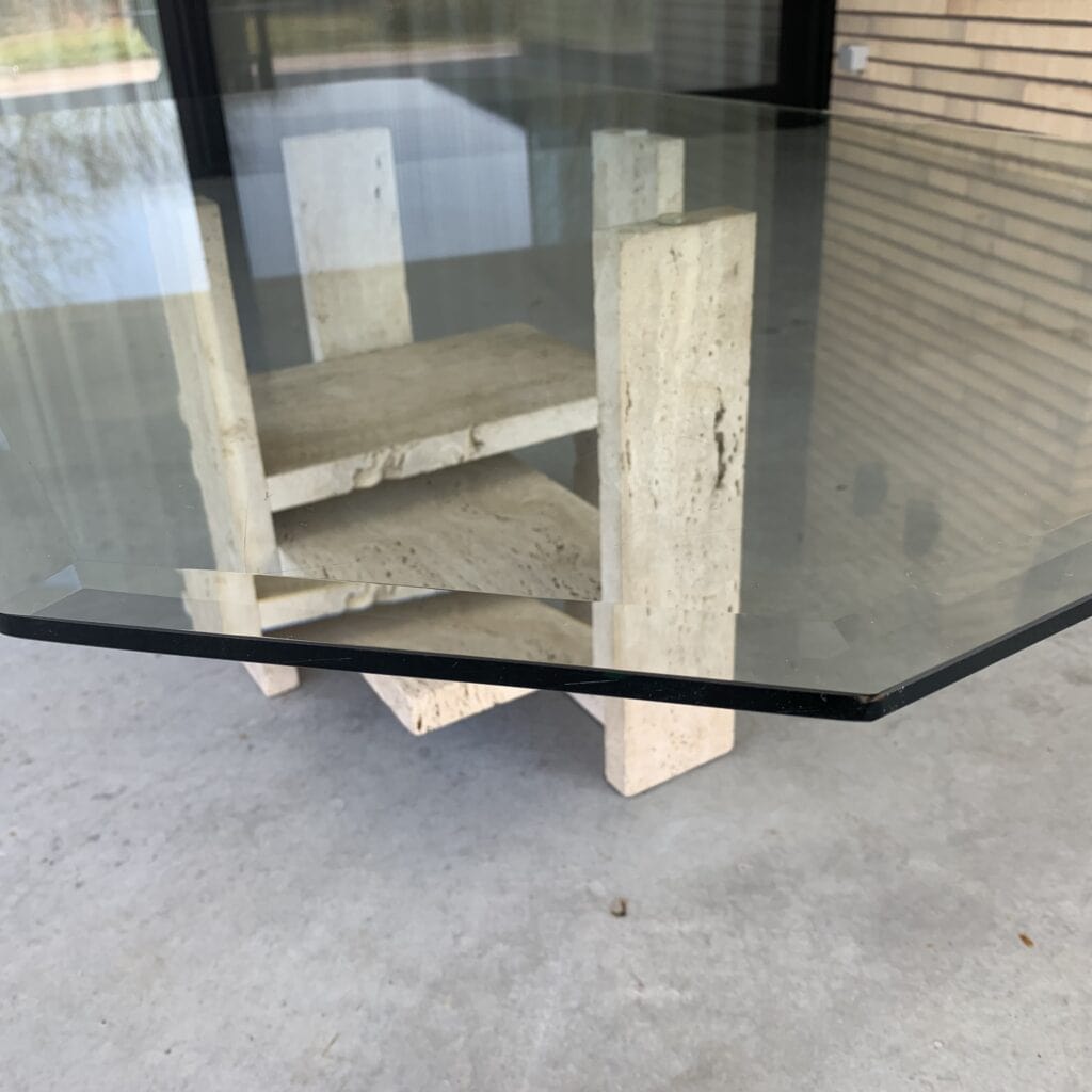 Een close-up van een glazen tafel met een gebarsten rand. De tafel heeft een stenen of betonnen voet met verticale en horizontale platen, en op de achtergrond zijn een groot raam en een bakstenen muur te zien.