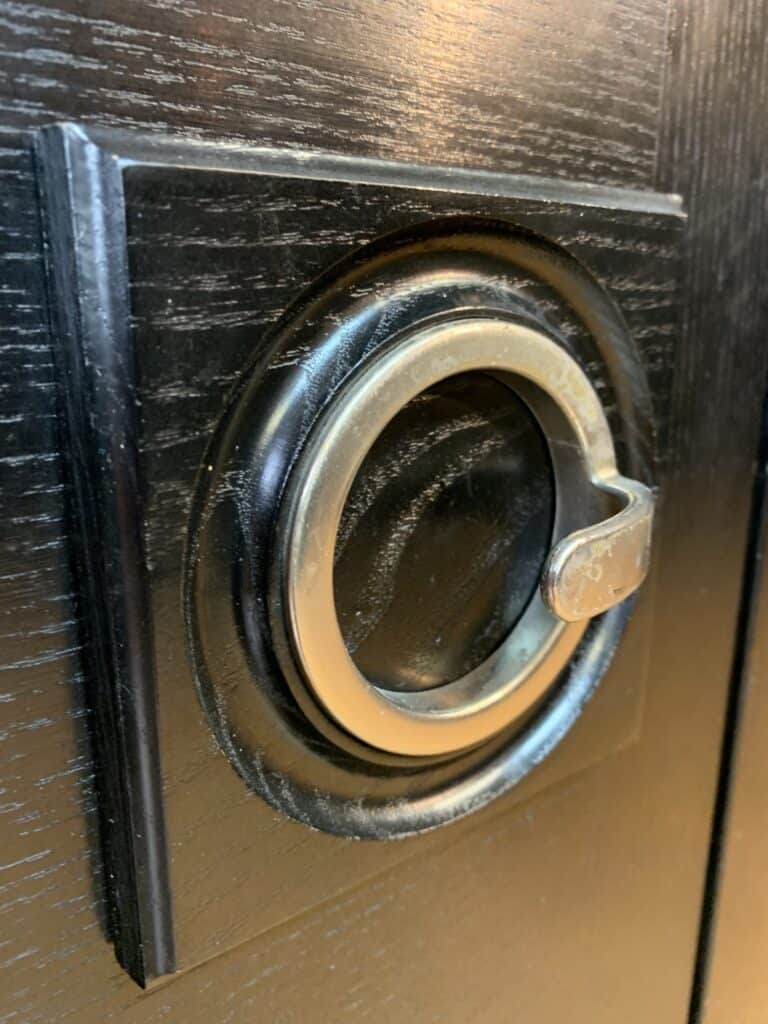 Close-up van een ronde messing handgreep met een grendel op een donker houten oppervlak, mogelijk een kast- of meubeldeur. Het hout heeft een zichtbare nerfstructuur en een glanzende afwerking.
