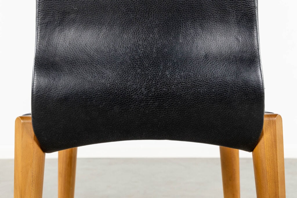 Close-up van de rugleuning van een stoel met zwarte leren bekleding en lichtgekleurde houten poten, tegen een effen witte achtergrond.