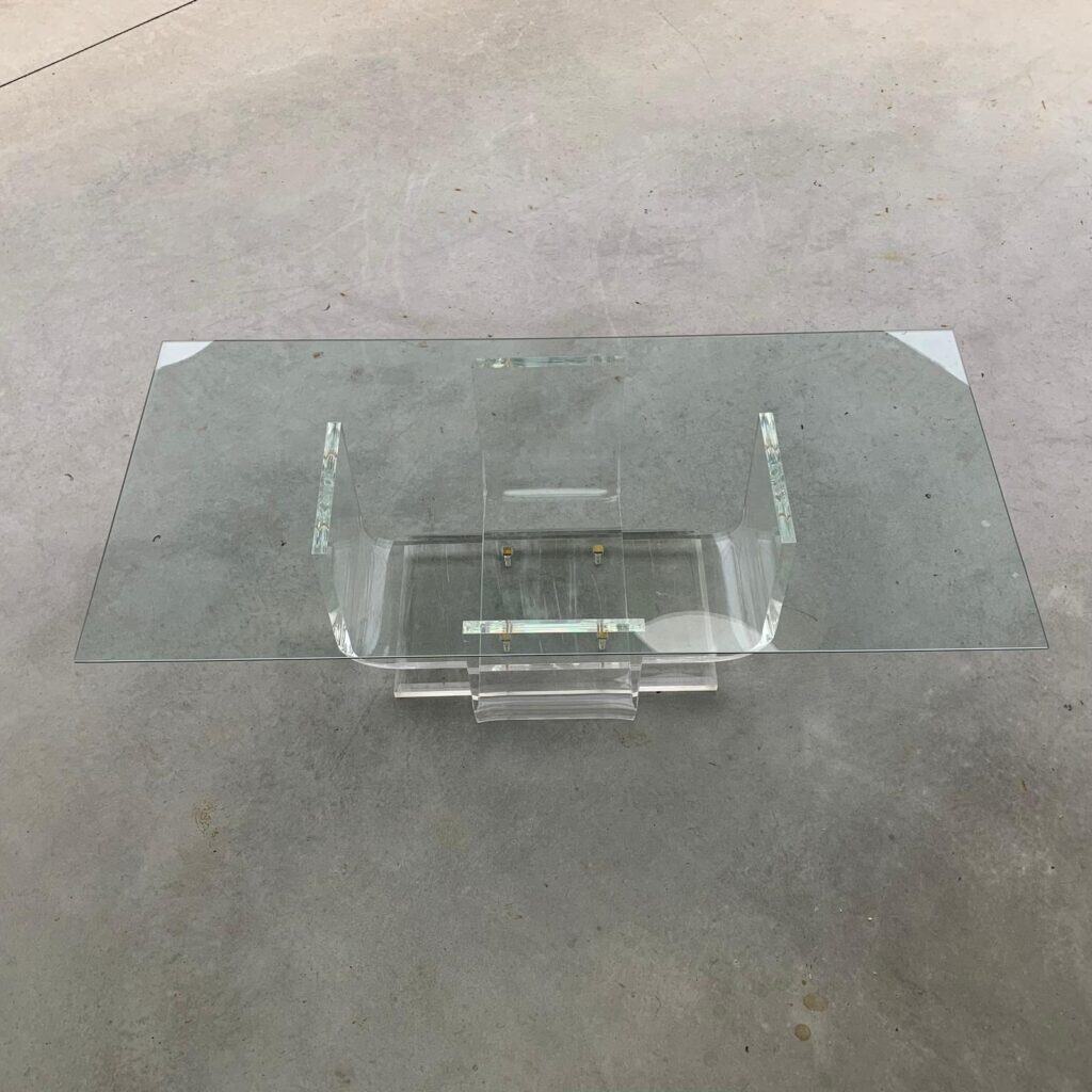 Een moderne rechthoekige glazen salontafel met een helder acryl onderstel, geplaatst op een gladde betonnen vloer. Het tafelblad heeft scherpe hoeken en het onderstel is voorzien van hoekige, transparante steunen.
