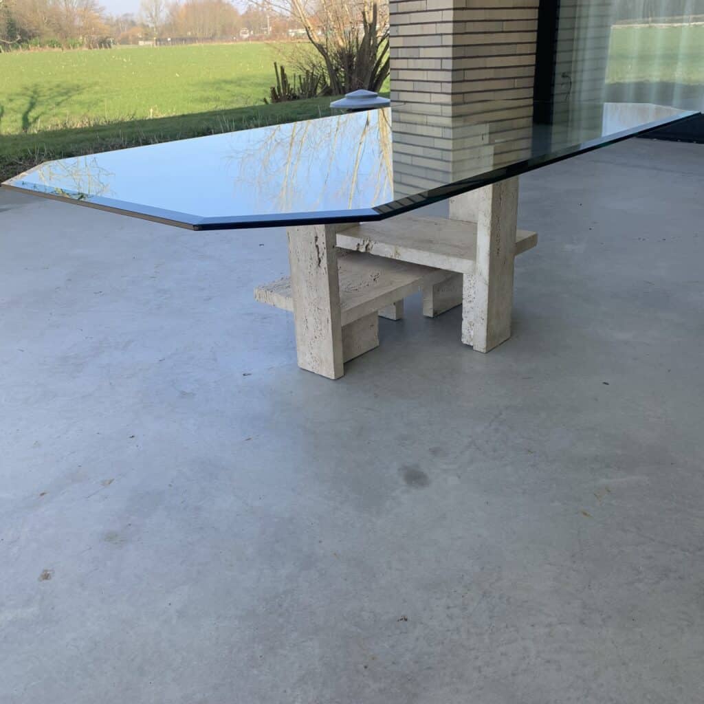 Een moderne glazen tafel met een geometrisch stenen onderstel staat op een betonnen terras. Op de achtergrond zijn groen gras en bomen zichtbaar, naast een bakstenen gebouw.