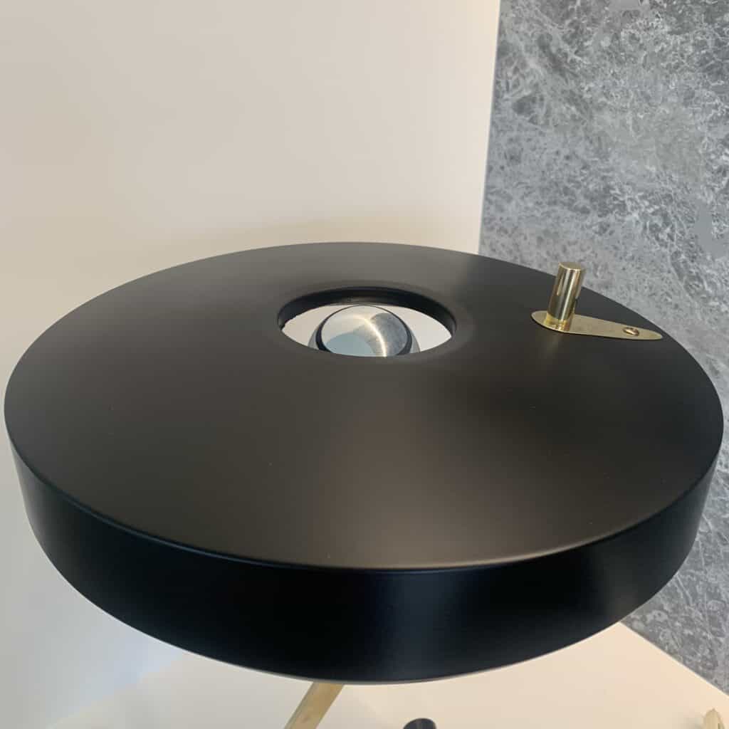 Een moderne, zwarte bureaulamp met een ronde kap, een metalen gloeilamp in het midden die zichtbaar is door een opening en een kleine messing schakelaar bovenop, tegen een tweekleurige achtergrond van wit en grijs marmer.