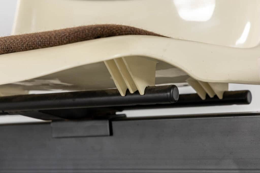 Close-up van de onderkant van een crèmekleurige plastic stoelzitting met een bruin kussen. De ondersteunende structuur en een zwarte metalen staaf daaronder zijn zichtbaar.