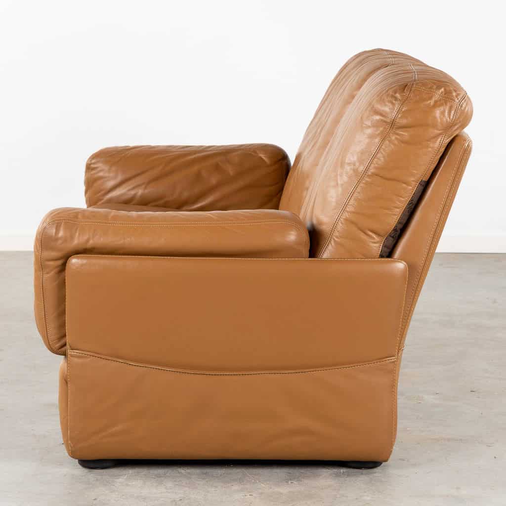 Zijaanzicht van een bruine leren fauteuil met een beklede rugleuning en zitting, geplaatst op een gladde betonnen vloer tegen een witte muur. De stoel heeft een modern, hoekig ontwerp met ronde, zachte armleuningen.