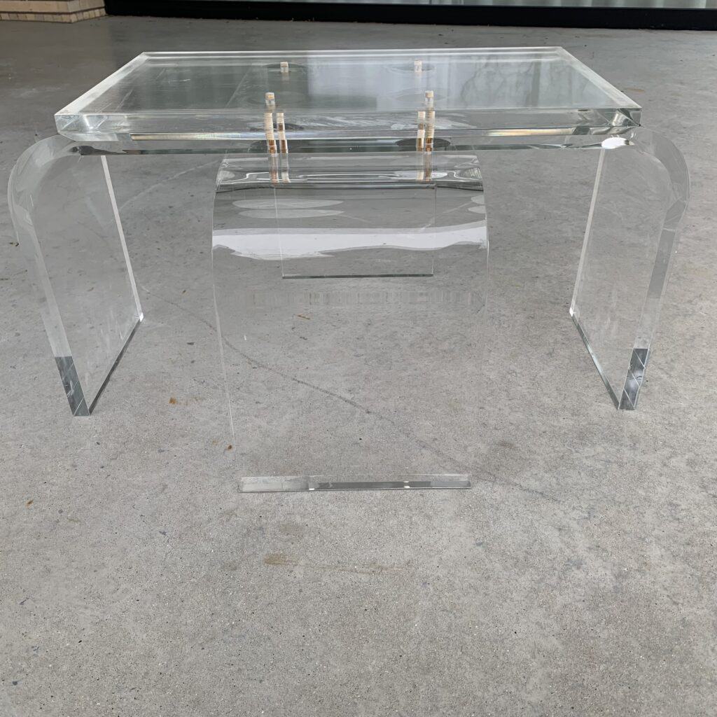 Een tafel van helder acryl of glas met een gebogen, modern design staat op een gladde, lichtgrijze betonnen vloer. De tafel heeft een vlak blad en vlakke zijpanelen, wat hem een minimalistische uitstraling geeft.