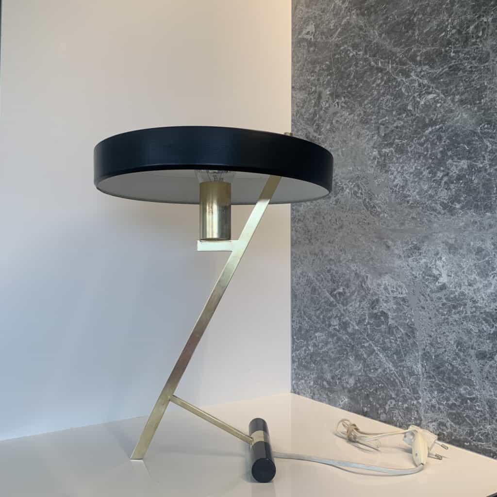 Een moderne tafellamp met een zwarte ronde kap, een hoekige metalen voet van goud en een wit snoer, geplaatst op een wit oppervlak naast een grijze marmeren muur.
