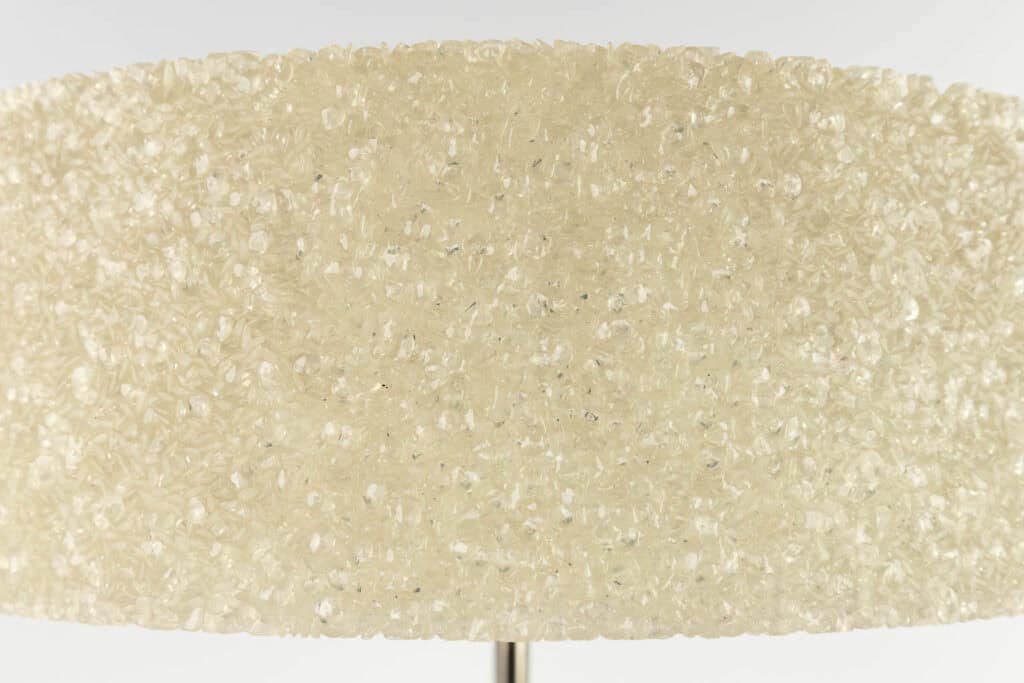 Close-up van een lampenkap bedekt met doorschijnende, getextureerde hars of kristalachtige kralen, waardoor deze een glinsterende, decoratieve uitstraling krijgt. De achtergrond is effen en lichtgekleurd.