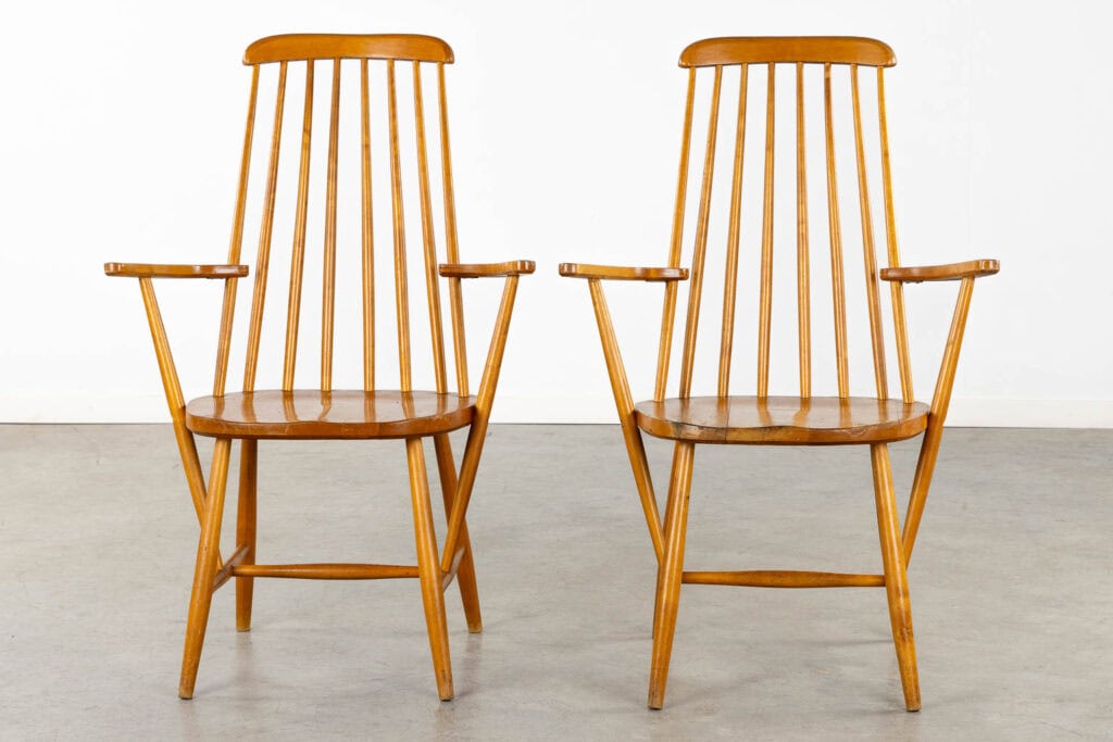 Twee houten fauteuils met hoge, smalle, spijlenvormige rugleuningen en schuine armleuningen, naast elkaar geplaatst op een grijze betonnen vloer tegen een eenvoudige witte muur.