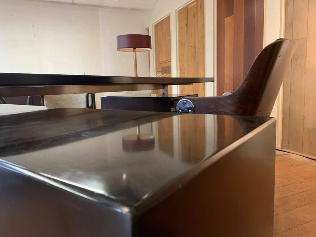 Een modern kantoor, genomen vanaf een laag standpunt, met een glanzend zwart bureau, een houten stoel met donkere armleuningen, een lamp met een kastanjebruine kap en houten lambrisering aan de muren op de achtergrond.