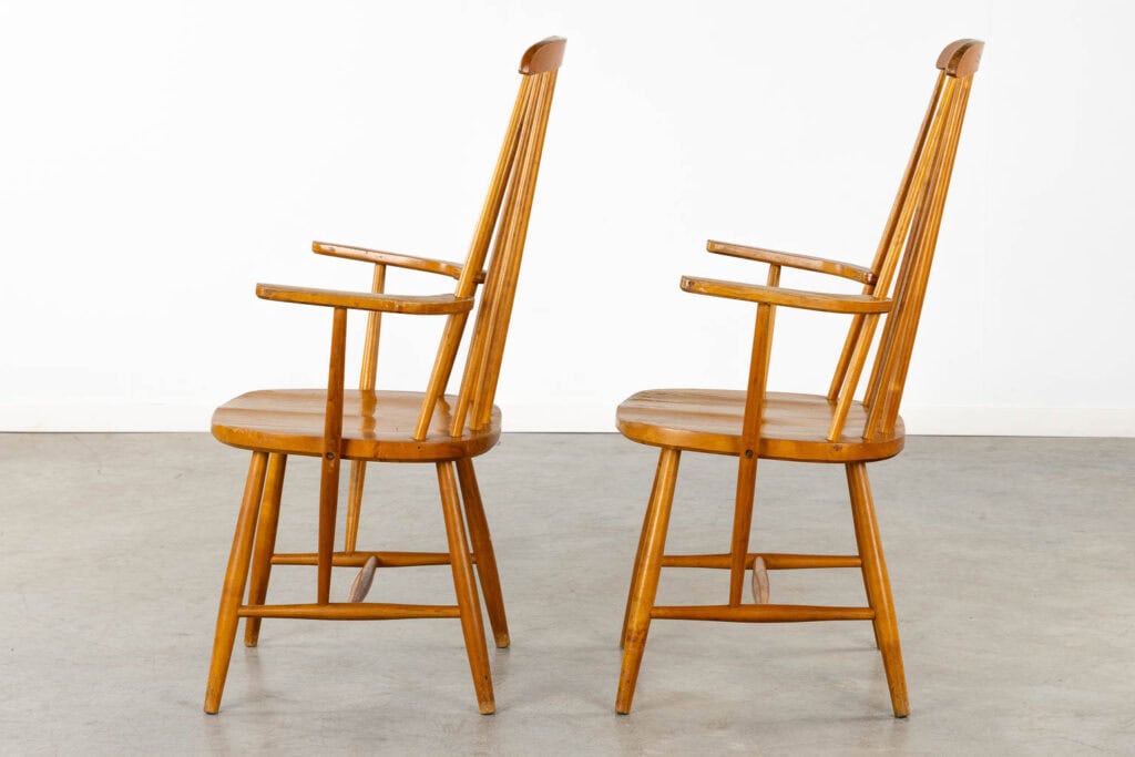 Twee houten fauteuils met spijlenleuningen en schuine poten staan naast elkaar, van opzij gezien, op een grijze betonnen vloer tegen een eenvoudige witte muur.