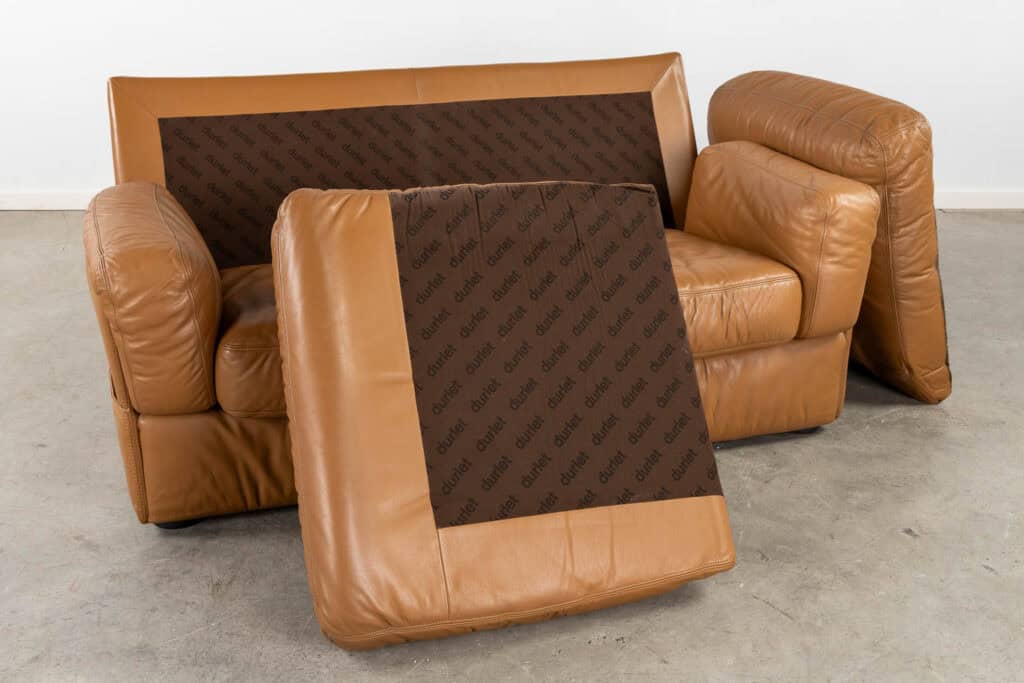 Een beige leren tweezitsbank waarvan de zit- en rugkussens verwijderd zijn en vooraan zijn geplaatst. De zichtbare kussenvoeten zijn bekleed met bruine stof met een herhalende tekst die lijkt te zeggen: 