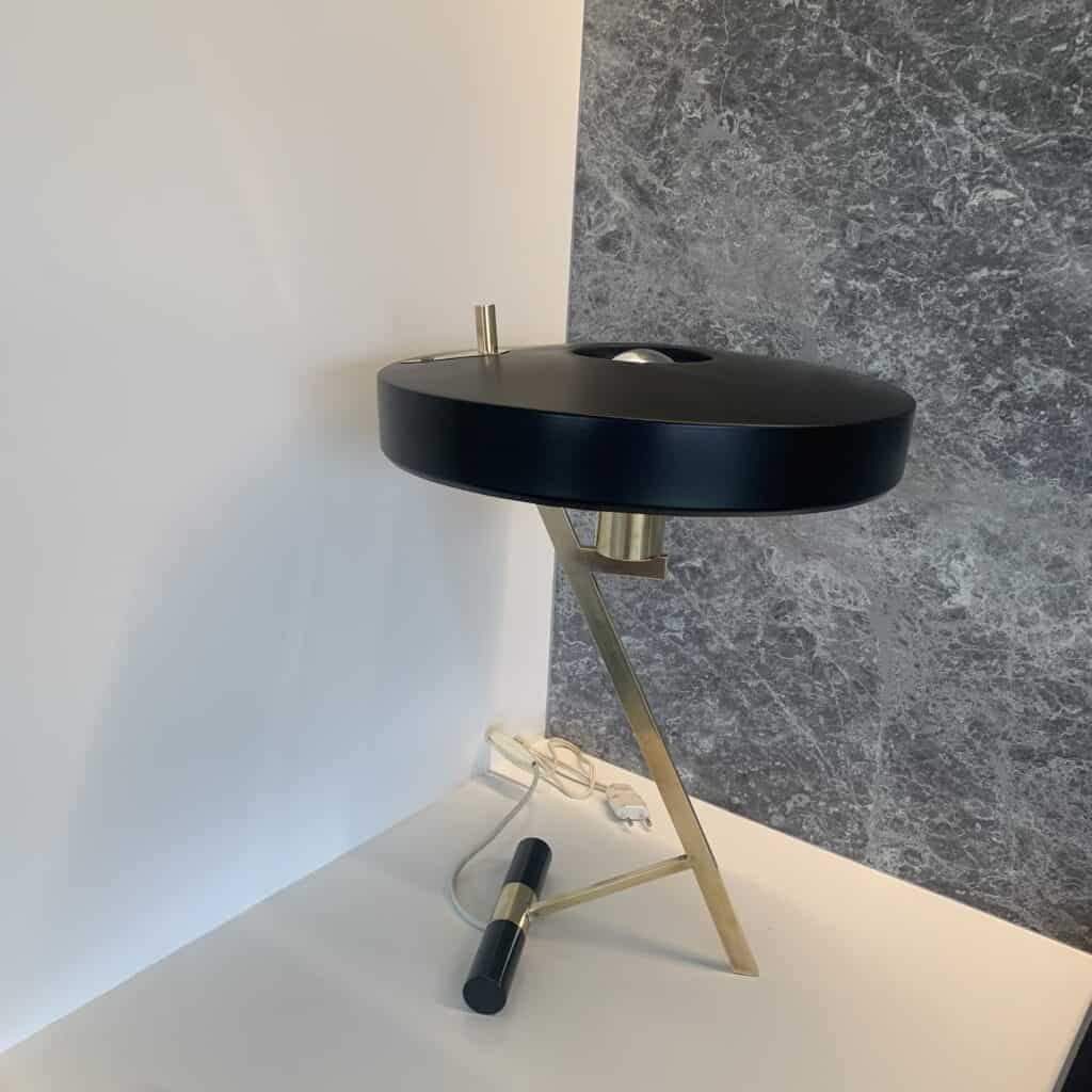Een moderne tafellamp met een ronde zwarte kap, een gouden hoekige voet en een zwarte cilindrische voet staat op een wit oppervlak naast een grijze muur met marmerstructuur. Achter de lamp is een snoer zichtbaar.