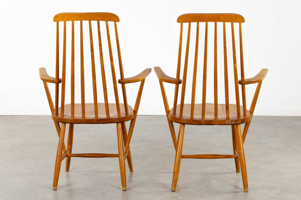 Twee houten fauteuils met hoge, spijlenvormige rugleuningen, schuine armleuningen en eenvoudige poten, naast elkaar geplaatst op een grijze vloer tegen een witte muur.